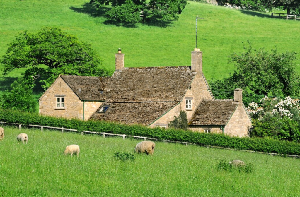 A stone cottage in lush countryside, with sheep grazing in the foreground and rolling hills beyond