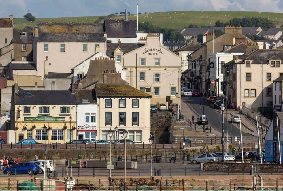 Coastal town with historic buildings, cars, pedestrians, and boats, with hills in the background