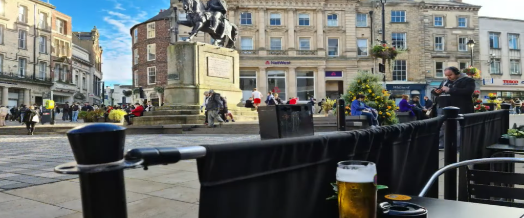 Bustling square with a statue, people, a street musician, and a foreground view of a cold beer